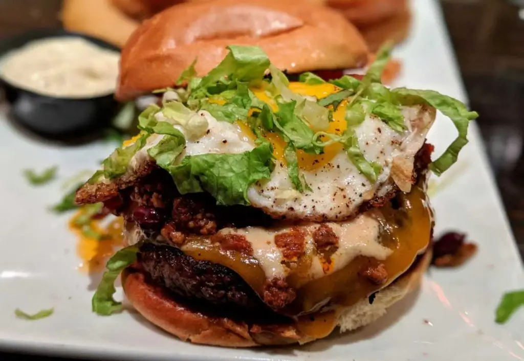 Kitchen Sink. H&F brioche bun, bacon, fried egg, chili, cheddar cheese, lettuce, tomato, bourbon, smoked tomato aioli. Onion rings.