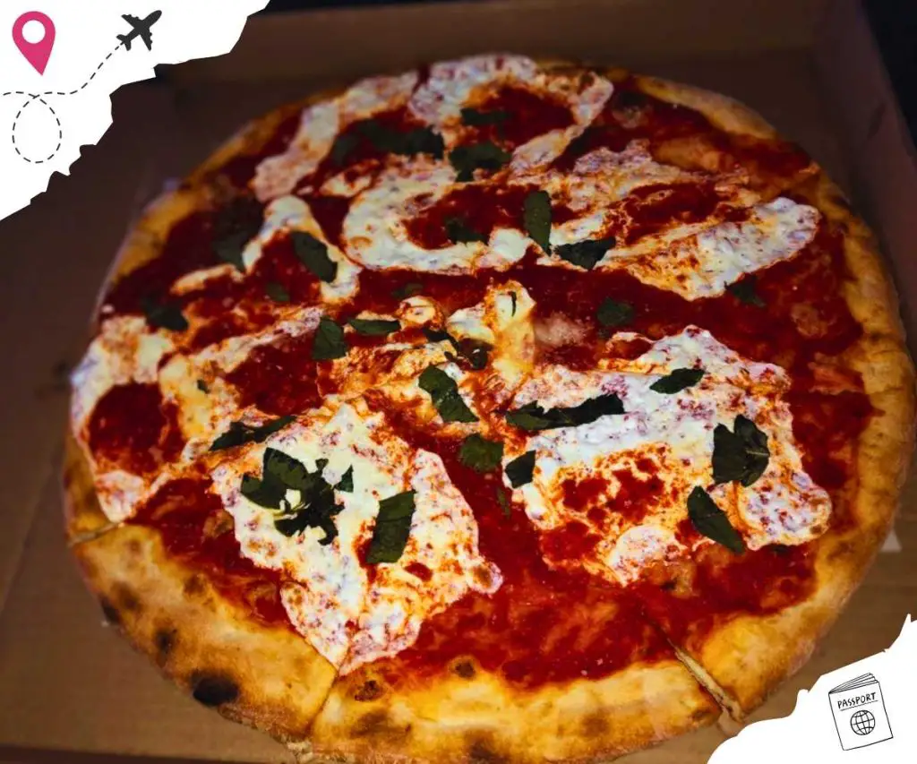 A pizza in a box topped with large white blotches of cheese and shreds of basil over tomato sauce.