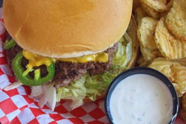 Crispy cheeseburger with lettuce, jalapenos, and creamy dipping sauce on red checkered paper - perfect for travel food lovers.