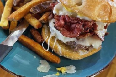 French fries with seasoned potato wedges served on a teal plate alongside a hearty bacon cheeseburger with a glazed glazed donut bun. The burger contains melted cheese, crispy bacon, and a beef patty.