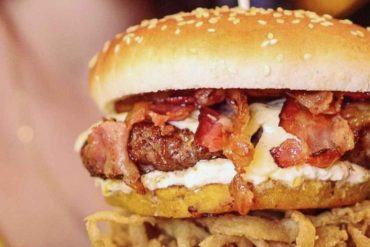 Delicious bacon cheeseburger with crispy fried onions, fresh toppings, and a small Colombian flag, served with a side of fries, highlighting American and international fast food cuisine.
