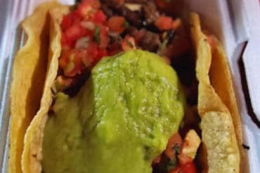 Fresh street-style taco with seasoned beef, fresh tomato salsa, and a generous scoop of vibrant green guacamole, served in a white takeout container. Perfect for authentic Mexican cuisine lovers and travelers exploring local food trends.