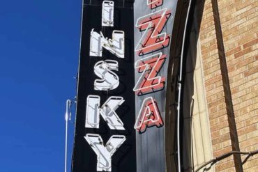 Food court restaurant neon sign with vintage style, illuminated words "KAYSS" and "PIZZA" against a bright blue sky. Perfect for travel and dining SEO keywords.