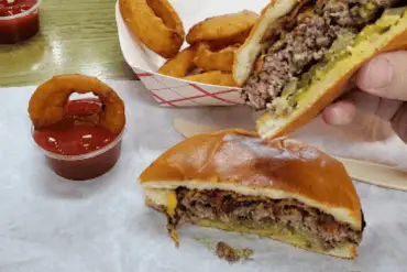 Cheeseburger with beef patty, pickles, and melted cheese on a soft bun, served with onion rings, french fries, and ketchup in a casual restaurant setting. Perfect comfort food for travel and food enthusiasts.