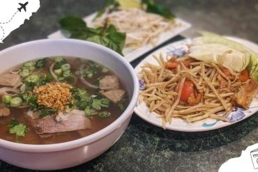 Steamed beef noodle soup with fresh herbs and fried garlic alongside a plate of stir-fried noodles with vegetables on a dark table. Authentic Asian cuisine highlighting flavors and traditional dishes.