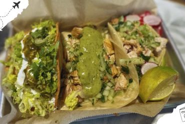Fresh Mexican tacos with green sauce, chopped vegetables, lime wedges, and savory fillings served in a paper tray, highlighting authentic street food experience and flavorful cuisine.