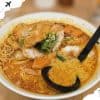 Crispy fried fish sitting atop a flavorful bowl of spicy ramen with vegetables, green onions, and a rich, fiery broth, served with a black spoon for an authentic Asian culinary experience.