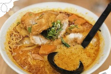 Crispy fried fish sitting atop a flavorful bowl of spicy ramen with vegetables, green onions, and a rich, fiery broth, served with a black spoon for an authentic Asian culinary experience.