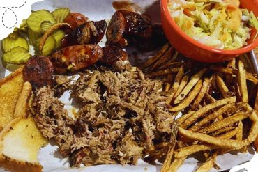 Succulent barbecue platter with pulled pork, crispy fries, grilled chicken wings, pickles, and fresh coleslaw, served on a wooden tray for a delicious American-style meal.