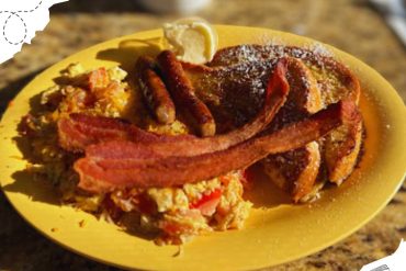 Savory breakfast plate with bacon, eggs, sausage links, and toast on a yellow plate in a rustic setting.