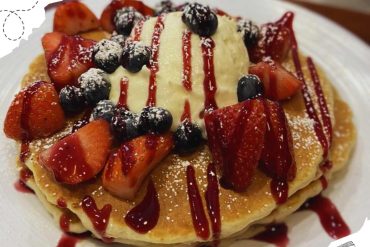 Fresh strawberry and blueberry pancake topped with vanilla ice cream, strawberry sauce, and powdered sugar, served on a white plate for a delicious breakfast or dessert.