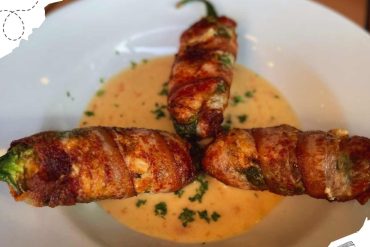 Succulent bacon-wrapped jalapeño poppers on a white plate with creamy dipping sauce, garnished with herbs, showcasing delicious international cuisine.