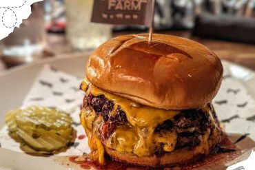 Beef burger with cheddar cheese, pickles, and melted toppings on a plate, served with a pickle spear and a small container of relish, at an restaurant or diner.