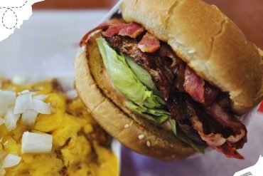 Smoked brisket sandwich with crispy bacon, fresh lettuce, and toasted bread served with a side of cheesy scrambled eggs topped with chopped onions, showcasing delicious American comfort food.
