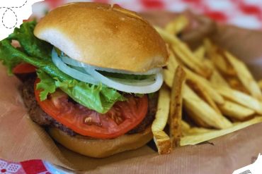 Burger with lettuce, tomato, onion, and beef patty served with French fries on a basket at a casual dining restaurant.