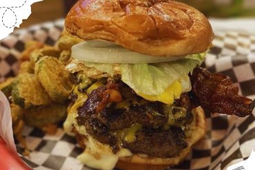 Juicy double cheeseburger with bacon, lettuce, onions, and crispy fried onion rings on a checkered paper tray, served at a restaurant or fast food place for American cuisine lovers.
