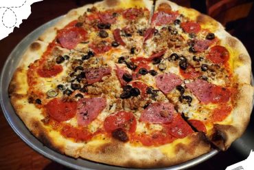 Pizza with pepperoni, sausage, black olives, and ham on a metal tray, served in a restaurant setting, showcasing delicious toppings and a crispy crust for a savory travel destination experience.