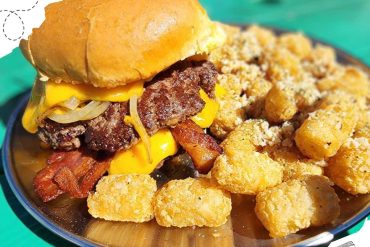 Juicy cheeseburger with crispy bacon, melted cheese, and fresh onions, served with golden tater tots on a stainless steel plate at a travel-themed eatery.