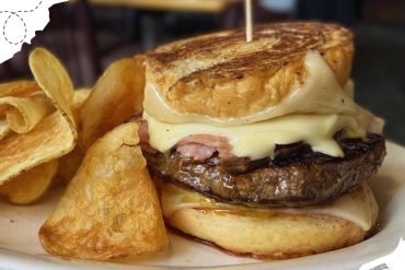 Fluffy breakfast sandwich with melted cheese, sausage patty, egg, and toasted bread, served with crispy potato chips on a white plate.