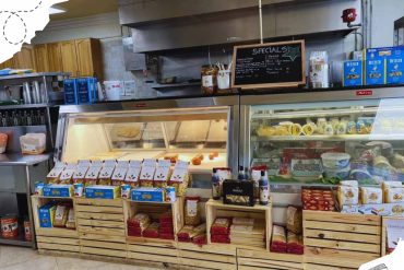 Organic pasta and cheese products displayed on wooden crates at a local Italian deli or grocery store, with fresh deli items, boxed products, and refrigerated goods behind the counter.