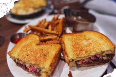 Golden toasted grilled cheese sandwich with roast beef, crispy fries, and dipping sauces on a white plate at a cozy restaurant, showcasing American comfort food and casual dining experiences.