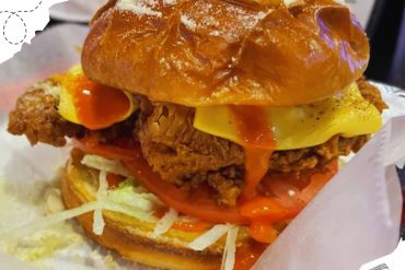 Crispy fried chicken sandwich with melted cheese, fresh lettuce, tomato, and spicy sauce on a shiny bun, served with shredded cheese and shredded lettuce underneath, delicious comfort food option.
