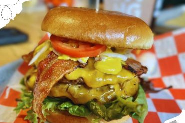 Bacon cheeseburger with fresh lettuce, tomato, crispy bacon, melted cheese, and a soft bun, served on checkered paper at a casual dining restaurant. Perfect for food lovers and burger enthusiasts.