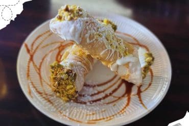 Cream-filled pastry topped with pistachios on a decorative plate, drizzled with caramel and white chocolate, showcasing delicious dessert options for travel and culinary experiences.