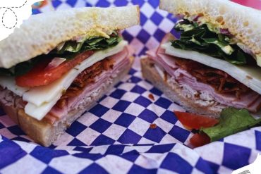 Crispy breakfast club sandwich with fresh vegetables and deli meats on checkered blue and white paper, ideal for travel and food lovers, showcasing delicious and portable meal options for destinations.