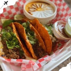 Crispy beef tacos with fresh cilantro and lime wedges, served with salsa and sauces, on red checkered paper tray - Mexican street food, authentic flavors, delicious and savory meal, vibrant presentation.