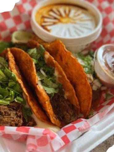 Crispy beef tacos with fresh cilantro and lime wedges, served with salsa and sauces, on red checkered paper tray - Mexican street food, authentic flavors, delicious and savory meal, vibrant presentation.