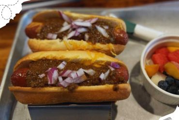Hot dog with chili, onions, and mustard served on a tray with a side of fresh fruit salad, popular street food in destination travel, delicious outdoor dining experience, Go To Destinations travel guide.