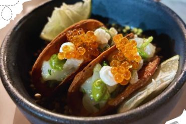 Fresh fish tacos topped with orange salmon roe, chopped onions, and lime wedges served in a black bowl, showcasing authentic Mexican cuisine and vibrant flavors.