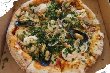 Fresh seafood pizza with clams, shrimp, mussels, and herbs, served in a takeout box, showcasing a delicious travel-inspired meal perfect for food lovers exploring new destinations.