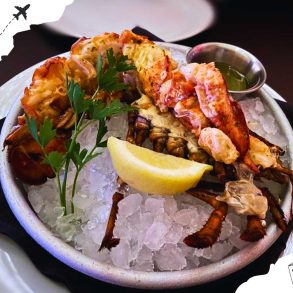 Dressed lobster with a cup of oil on a bowl of ice, garnished with a lemon slice and parsley snippets.