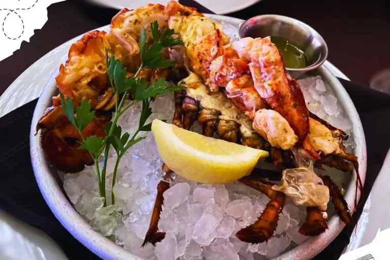 Dressed lobster with a cup of oil on a bowl of ice, garnished with a lemon slice and parsley snippets.