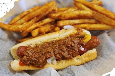 Hot dog with chili and fries, classic American fast food served on parchment paper.