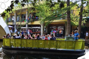 Riverboat cruise with tourists enjoying a scenic ride through a charming urban area surrounded by trees and colorful buildings, perfect for sightseeing and exploring city attractions.