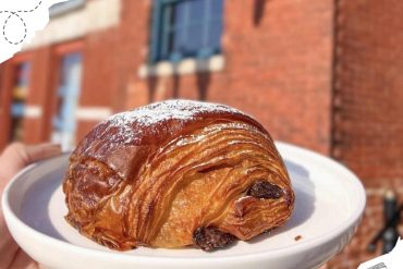 Crusty cinnamon roll pastry with raisins on a white plate outdoors with a red brick building in the background.