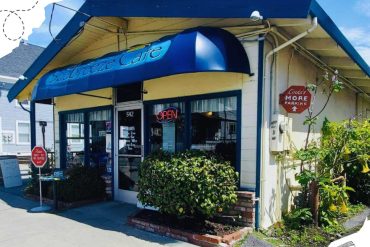 Gulf Breeze Cafe exterior with blue awning, open sign, surrounding greenery, and clear sky in a charming coastal town setting. Ideal for travel destinations and sightseeing.