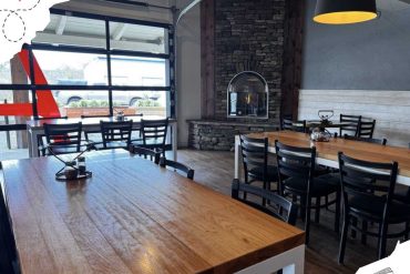 Modern cozy restaurant interior with wooden tables, black chairs, stone fireplace, and large windows offering natural light, ideal for a comfortable travel dining experience.