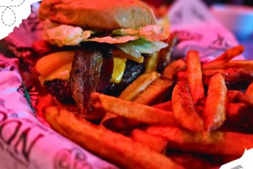 Juicy gourmet burger with crispy fries, melted cheese, bacon, lettuce, and pickles on a soft bun at a lively dining spot. Perfect comfort food for a travel food adventure.