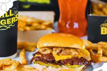 A grilled onion smashburger surrounded in scattered French fries and drinks.