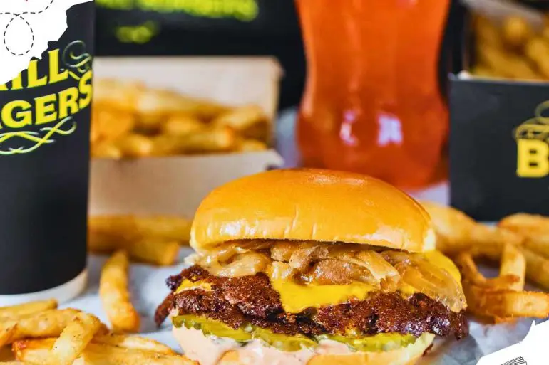 A grilled onion smashburger surrounded in scattered French fries and drinks.