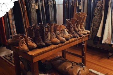 Worn leather boots and men's trousers on display at a thrift store or clothing shop, featuring brown and tan hiking footwear, casual pants, and a variety of accessories for travel or outdoor adventures.