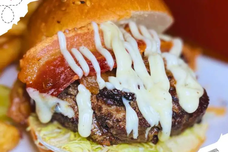 Burger with the top bun lopsided, revealing bacon and a beef patty drizzled with creamy sauce.