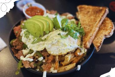 Savory Mexican carne asada fries bowl topped with lettuce, guacamole, fried egg, and crispy toasted bread slices, served with spicy salsa on the side, perfect for food travel experiences.