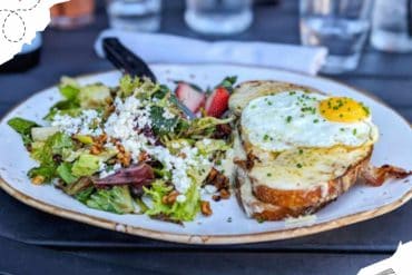 Fresh breakfast plate featuring a sunny-side-up egg on toasted bread with chopped chives, paired with a colorful mixed greens salad topped with crumbled cheese, strawberries, and nuts at an outdoor cafe.