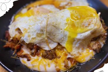 Savory breakfast dish with melted cheese, seasoned meats, and fried egg on top in a black skillet, popular comfort food, delicious brunch option, hearty meal, served hot.