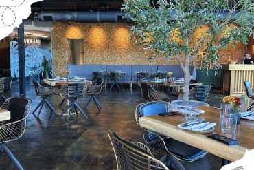 Modern restaurant interior with stylish metal chairs, wooden tables, a large leafy tree decor, and warm ambient lighting, perfect for travel and culinary destination experiences.